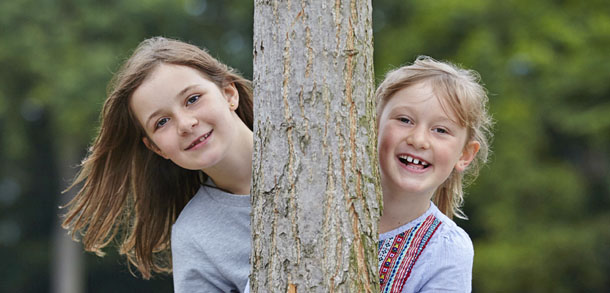 Sisters peer out from behind a tree-trunk, perhaps playing hide and seek